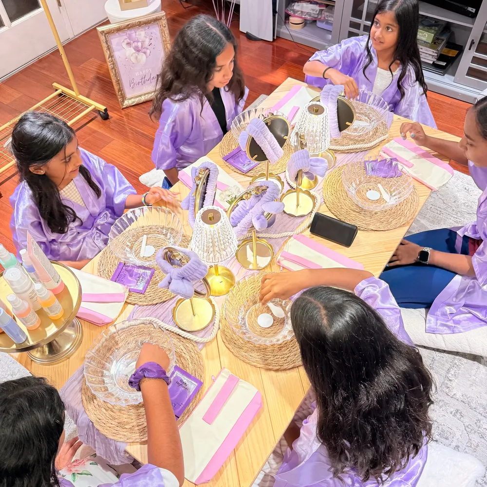 Guests in lavender robes make crafts together at a decorated spa party table.
